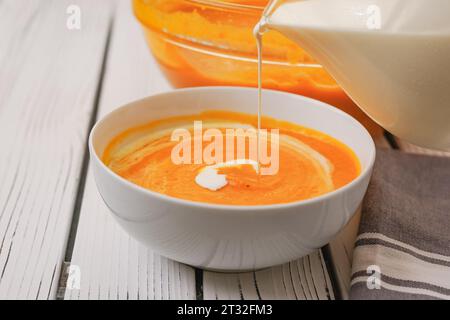 Soupe de courge musquée rôtie servie avec un bacon croustillant et une garniture épaisse à la crème, gros plan dans un bol sur une planche de bois blanche Banque D'Images