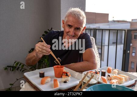 Attrayant homme d'âge moyen mangeant assis à une table posée avec de la nourriture chinoise à emporter sur un balcon de la ville Banque D'Images