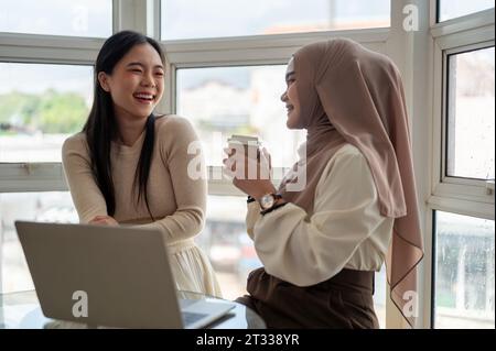 Une jeune femme asiatique joyeuse et belle aime parler avec son amie musulmane, rire, s'amuser dans un café. Concept de style de vie Banque D'Images