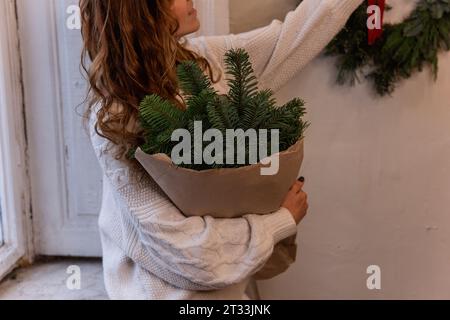 Portrait sans visage de jeune femme en pull tricoté blanc, assis sur le rebord de la fenêtre, tenant dans les mains bouquet de Noël en papier artisanal près de la fenêtre. ECO avec Banque D'Images