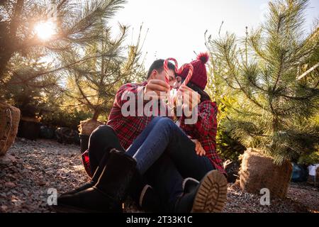 La canne à bonbons blanc rouge est tenue dans les mains tendues d'un couple amoureux dans des chemises à carreaux, des chapeaux tricotés près du marché vert des arbres de Noël. Homme heureux Banque D'Images