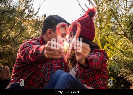 Couple amoureux en chemises à carreaux, chapeaux tricotés tiennent des cannes de bonbons en forme de cœur dans les mains près du marché vert des arbres de Noël. L'homme et la femme s'embrassent, rient, h. Banque D'Images