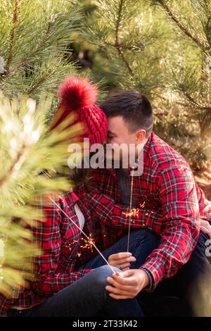 Joyeux jeune couple dans des chemises à carreaux rouges s'embrassant, tenant des étincelles festives dans les mains dans la forêt. Homme et femme rient au marché des arbres de Noël. CLO Banque D'Images