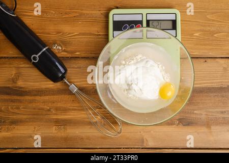 Farine, œuf, eau et crème sure dans un bol sur une balance de cuisine, vue de dessus Banque D'Images