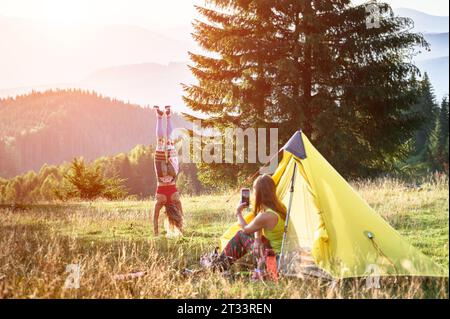 Deux femmes randonneurs campant en plein air. Jeune femme voyageant en montagne, randonnée en été. Concentrez-vous sur une femme sportive faisant du salto, une autre femme assise, prenant des photos. Concept de tourisme et d'aventure. Banque D'Images