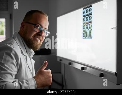 Vue latérale du médecin portant des lunettes examinant le scanner des poumons. Spécialiste masculin portant une robe blanche, regardant la caméra, souriant, montrant super. Concept d'hôpital et de santé. Banque D'Images