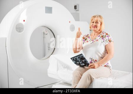 Tomodensitométrie médicale ou scanner IRM. Portrait d'une patiente en IRM. Femme blonde tenant le résultat d'IRM, scanner, souriant à la caméra, montrant les pouces vers le haut. Concept de médecine et de santé. Banque D'Images