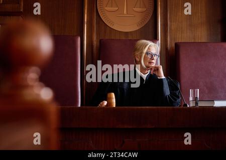 Juge blonde sérieuse dans le manteau noir frappant le marteau en bois sur le bloc sonore et prononçant le verdict ou le tribunal pendant le procès Banque D'Images