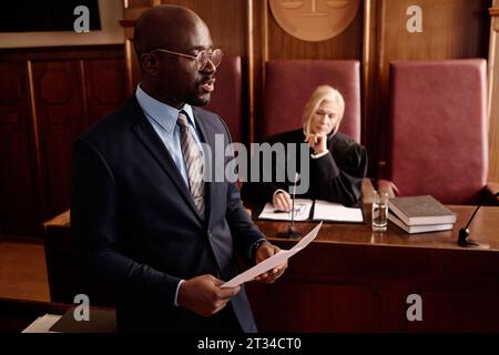 Jeune avocat confiant avec document juridique défendant le suspect tout en se tenant devant le côté de l'accusation et contre une femme juge mature Banque D'Images