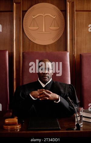 Juge masculin afro-américain mûr sérieux dans les lunettes et manteau noir regardant la caméra tout en étant assis par le bureau sur la chaise en cuir Banque D'Images