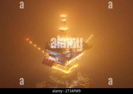 Grosse plate-forme pétrolière travaillant sur un océan ouvert pendant une nuit brumeuse. D'innombrables lumières illuminent la brume et créent un reflet dans les vagues d'eau. Symbole de Banque D'Images