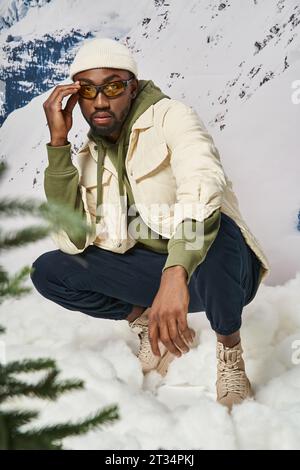 bel homme élégant dans une tenue chaude et modish accroupissant et touchant ses lunettes de soleil, concept d'hiver Banque D'Images
