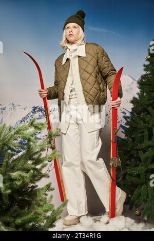 femme blonde élégante en chapeau de bobble et veste d'hiver avec des skis à côté des pins, concept d'hiver Banque D'Images