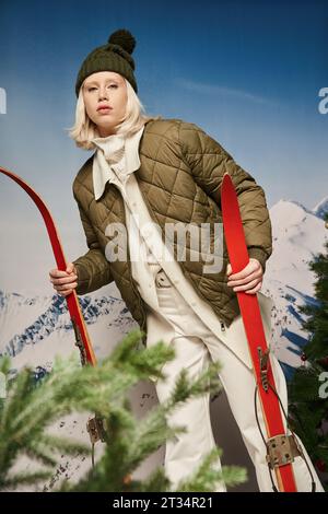 jolie jeune femme en veste chaude posant avec des skis sur fond enneigé, concept d'hiver Banque D'Images