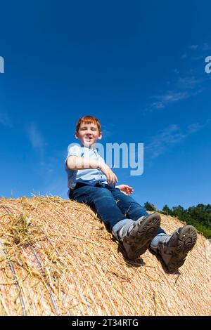 un garçon aux cheveux rouges assis sur une pile de paille dorée dans un champ, un garçon sur une pile de paille de blé épineux, un petit garçon mignon, portrait d'un garçon contre un bleu Banque D'Images