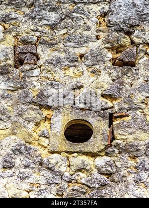 Petit trou de ventilation rond et extrémités de poutres en bois faisant un «visage drôle» dans le vieux mur de pierre - centre de la France. Banque D'Images