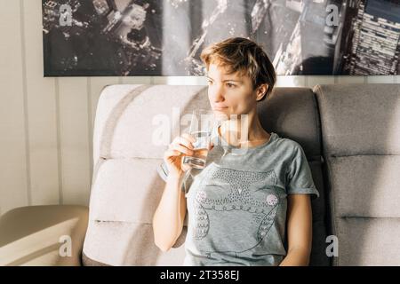 Une femme est assise sur un canapé au soleil et tient un verre d'eau Banque D'Images