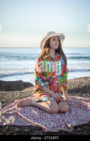 Dans la lueur de fin d'après-midi, une femme caucasienne de 20 ans est assise sur un rocher de plage, ornée d'une robe chemise vibrante à motifs et d'un chapeau chic Banque D'Images