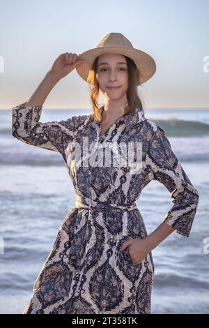 Une femme caucasienne de 20 ans se tient sur la plage au coucher du soleil, portant une robe chemise à motifs vibrante et colorée alors que le soleil projette une lueur chaude sur celle-ci Banque D'Images