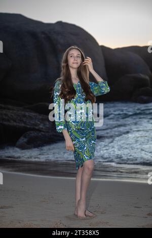 Une élégante femme caucasienne de 20 ans avec de longs cheveux fluides respire la grâce dans un plan rapproché, enfilée dans une robe chemise à motifs verts sur la plage Banque D'Images