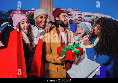 Groupe de jeunes adultes multiraciaux offrant un cadeau de noël à leur meilleur ami. Homme heureux recevant un cadeau surprise de Noël. Gars reconnaissant tenant une boîte emballée spéciale noel sur les vacances d'hiver. Photo de haute qualité Banque D'Images