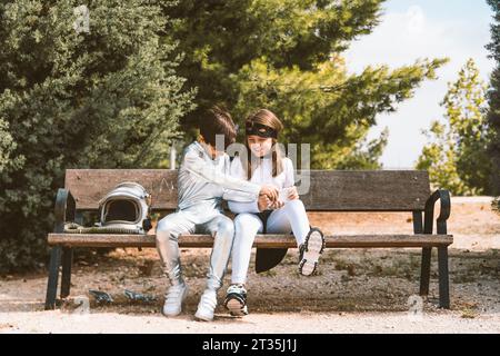 Deux enfants en costumes d'astronaute et de super-héros avec téléphone portable sur le banc du parc Banque D'Images