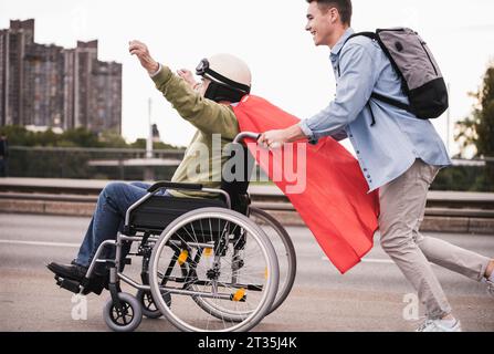 Jeune homme poussant des hauts homme assis dans un fauteuil roulant habillé en super héros Banque D'Images