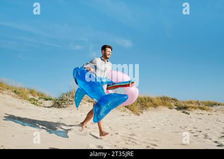Homme heureux courant avec anneau de natation gonflable et requin jouet à la plage le jour ensoleillé Banque D'Images
