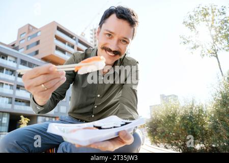 Homme d'affaires souriant tenant des sushis dans des baguettes assis au parc de bureaux Banque D'Images