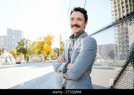 Homme d'affaires souriant confiant avec les bras croisés debout près de l'immeuble de bureaux moderne Banque D'Images