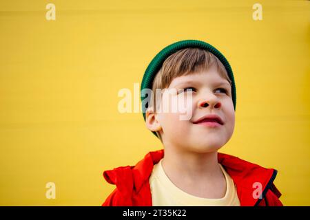 Garçon réfléchi portant une veste rouge et un chapeau en tricot devant le mur jaune Banque D'Images