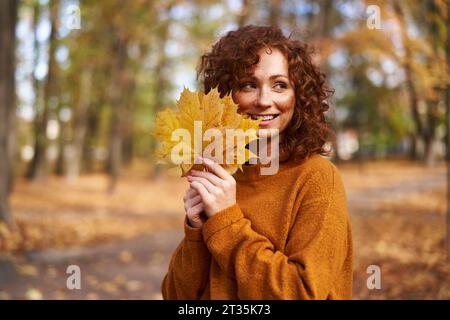 Femme rousse souriante tenant des feuilles d'érable au parc d'automne Banque D'Images