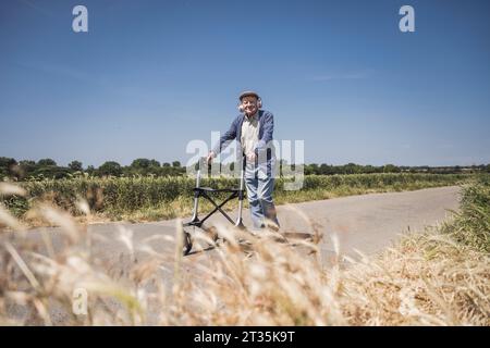 Homme âgé souriant portant des écouteurs sans fil marchant avec marcheur de mobilité sur la route Banque D'Images