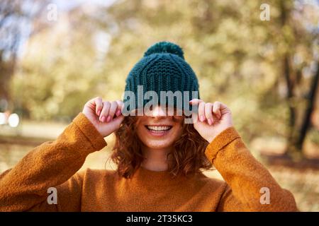 Joyeuse femme rousse couvrant les yeux avec un chapeau en tricot au parc d'automne Banque D'Images