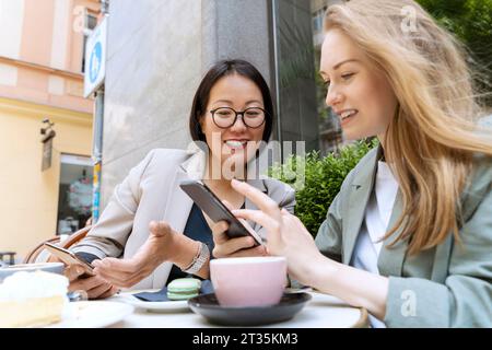 Heureux partenaires d'affaires discutant avec des téléphones intelligents assis au café Sidewalk Banque D'Images