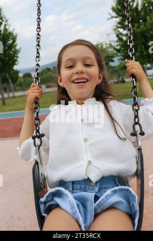 Fille heureuse assise sur la balançoire à l'aire de jeux Banque D'Images