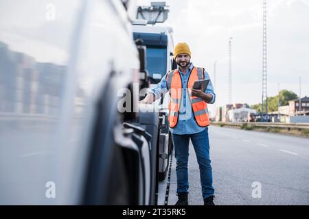 Chauffeur de camion souriant portant un chapeau de tricot debout avec tablette PC par camion Banque D'Images