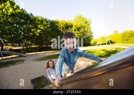 Fils souriant grimpant sur toboggan avec mère en arrière-plan Banque D'Images