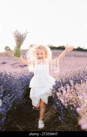 Fille blonde tenant bouquet de fleurs de lavande courant dans le champ Banque D'Images