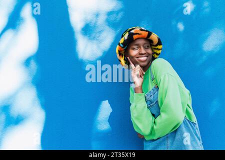 Heureuse jeune femme portant un chapeau coloré touchant le visage devant le mur bleu Banque D'Images