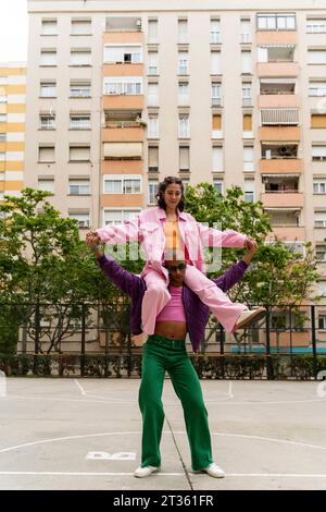 Personne non binaire tenant les mains d'une femme assise sur les épaules sur un terrain de basket-ball Banque D'Images