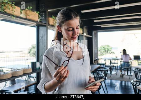 Portrait de jolie blonde debout dans un café avec des lunettes et un téléphone intelligent dans les mains Banque D'Images