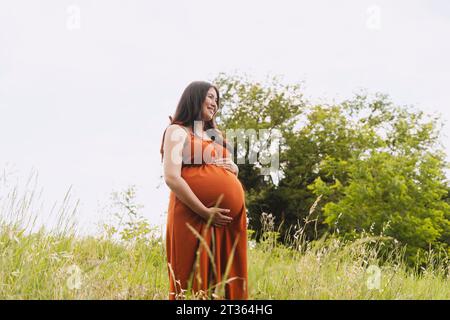 Femme enceinte souriante avec les mains sur le ventre debout au milieu de l'herbe Banque D'Images