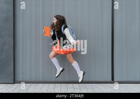 Écolière souriante tenant le livre et sautant devant le mur Banque D'Images