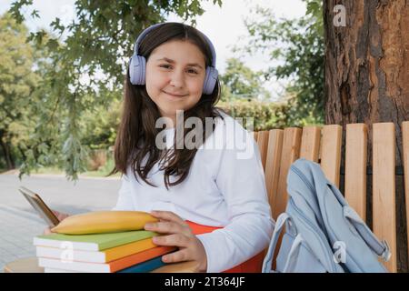 Écolière souriante avec des livres portant des écouteurs sans fil et assis sur un banc Banque D'Images