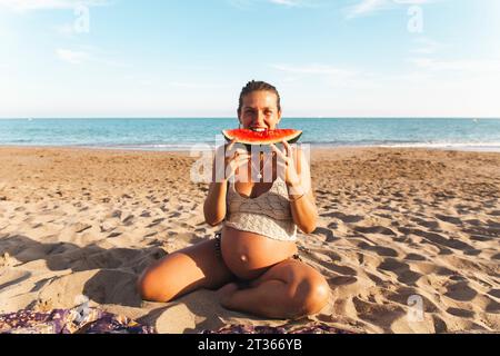 Femme enceinte assise sur le sable et mangeant de la pastèque à la plage Banque D'Images