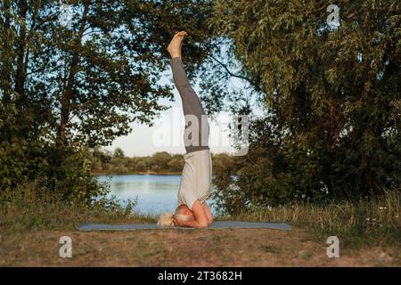 Femme mature pratiquant le yoga headstand sur le tapis au bord du lac Banque D'Images