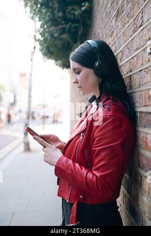 Femme portant des écouteurs sans fil à l'aide d'un téléphone intelligent près du mur Banque D'Images