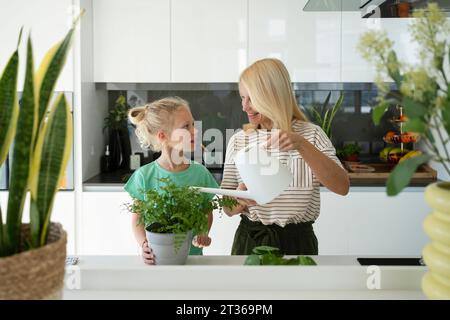 Femme heureuse arrosant la plante avec sa fille debout dans la cuisine à la maison Banque D'Images