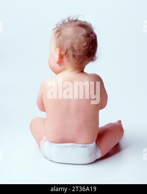 Bébé dans la couche jetable assis droit face à l'extérieur sur fond blanc. Banque D'Images